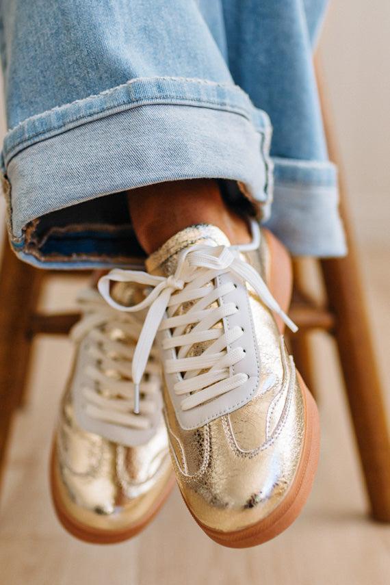 Woman wearing metallic gold and white slip-on sneakers featuring a bold swoosh pattern on the side, giving them a dynamic, sporty look. The durable gum sole provides excellent traction and a retro touch.