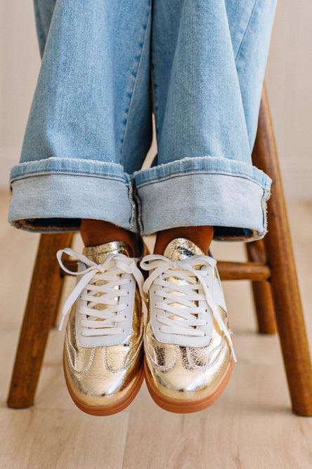 Woman wearing metallic gold and white slip-on sneakers featuring a bold swoosh pattern on the side, giving them a dynamic, sporty look. The durable gum sole provides excellent traction and a retro touch.