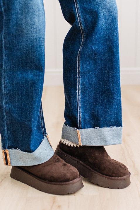 A close-up of a person's legs wearing blue cuffed jeans and dark brown suede platform boots on a light wood floor.