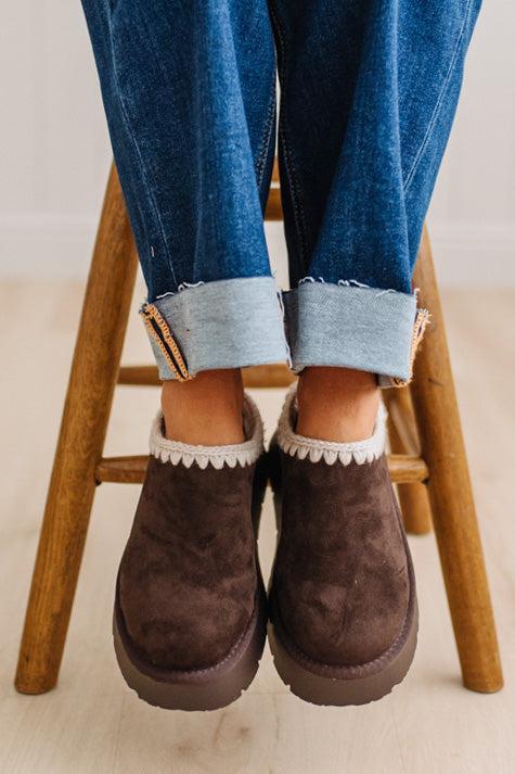 A person wearing brown suede slip-on slippers with white trim, paired with cuffed blue jeans, sitting on a wooden stool.