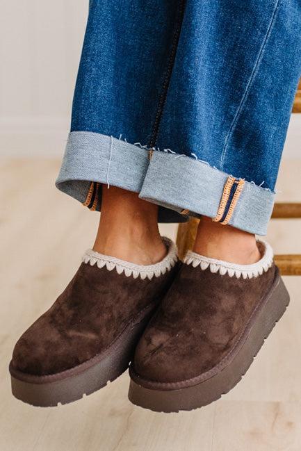 A person wearing dark brown suede slip-on slippers with a white scalloped trim and thick brown platform soles, paired with cuffed blue jeans on a light wood floor.