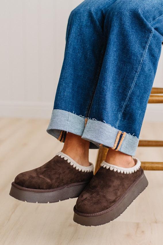 A person wearing brown suede slip-on clogs with white pom-pom trim and thick dark soles, paired with cuffed blue jeans, sitting on a wooden chair in a bright indoor setting.