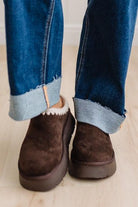 A close-up of a person's feet wearing dark brown suede slip-on boots with a white sherpa lining and platform soles, styled with cuffed blue jeans on a light wood floor.