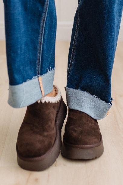 A close-up of a person's feet wearing dark brown suede slip-on boots with a white sherpa lining and platform soles, styled with cuffed blue jeans on a light wood floor.