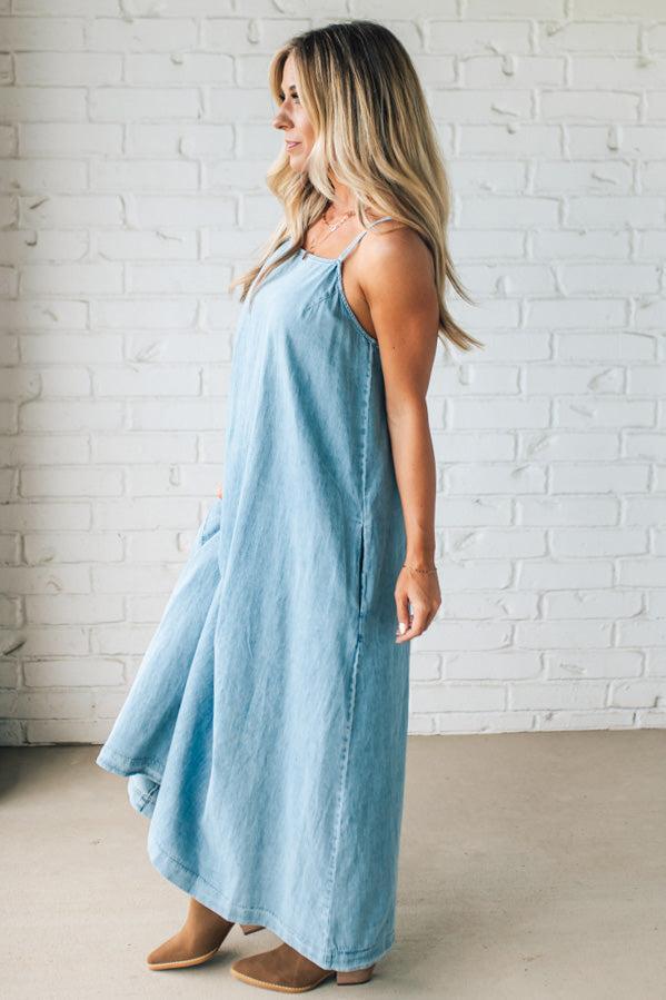 Blonde woman wearing a long denim maxi dress with adjustable spaghetti straps, side pockets, and a side slit.