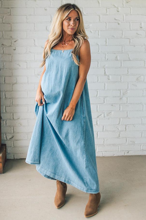 Blonde woman wearing a long denim maxi dress with adjustable spaghetti straps, side pockets, and a side slit.