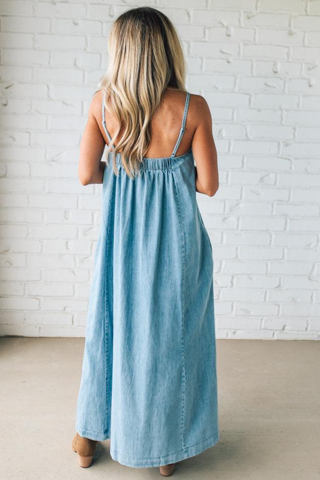 Blonde woman wearing a long denim maxi dress with adjustable spaghetti straps, side pockets, and a side slit.