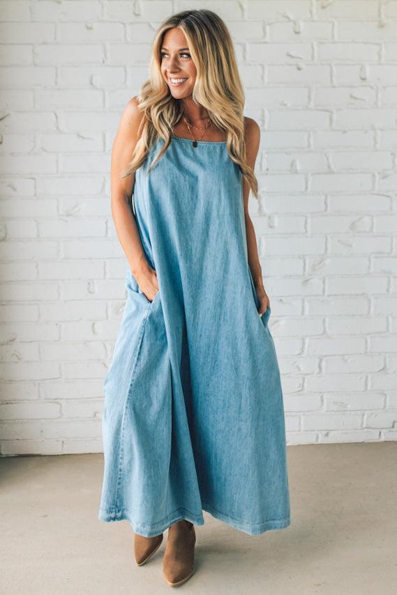 Blonde woman wearing a long denim maxi dress with adjustable spaghetti straps, side pockets, and a side slit.