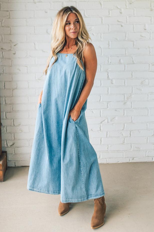 Blonde woman wearing a long denim maxi dress with adjustable spaghetti straps, side pockets, and a side slit.