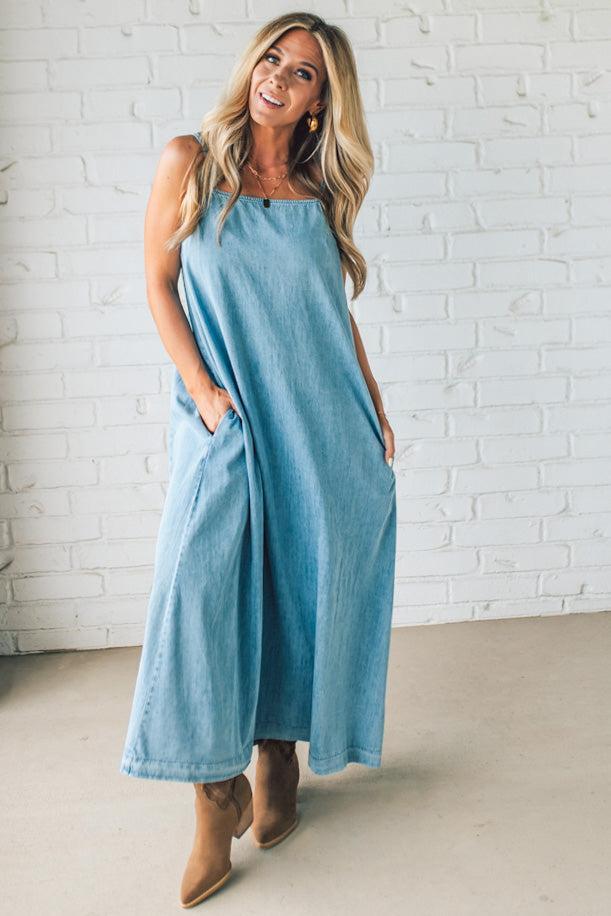 Blonde woman wearing a long denim maxi dress with adjustable spaghetti straps, side pockets, and a side slit.
