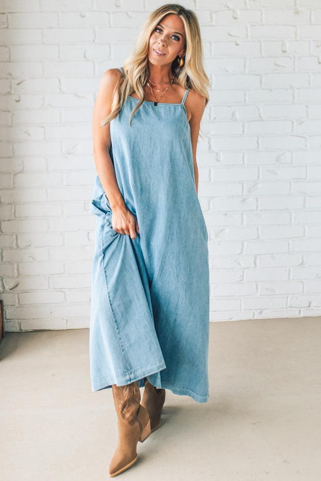 Blonde woman wearing a long denim maxi dress with adjustable spaghetti straps, side pockets, and a side slit.