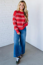 Woman wearing a red striped sweater and blue jeans standing against a white brick wall.