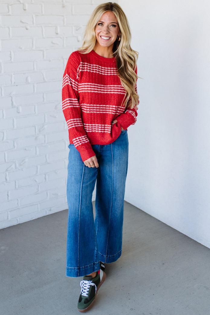 Woman wearing a red striped sweater and blue jeans standing against a white brick wall.