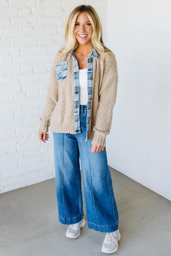 Woman wearing a beige cardigan, plaid shirt, blue jeans, and white sneakers against a white brick wall.
