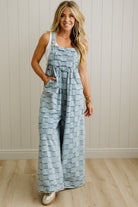 Woman wearing a blue patterned dress overalls against a white paneled wall.