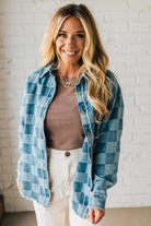 Woman wearing a blue plaid shirt over a brown top and white pants against a white brick wall.