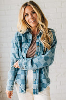 Woman wearing a blue plaid shirt over a brown top against a white brick wall.