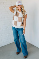 Woman wearing a checkered sleeveless top, blue jeans, and a white cap with a logo against a white brick wall.