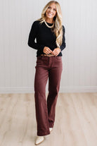 Blonde woman standing in a brightly lit studio with light wood floors and paneled wall behind. Wearing dark pants and a ribbed slim fit long sleeve sweater in black.