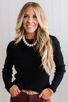 Blonde woman standing in a brightly lit studio with light wood floors and paneled wall behind. Wearing dark pants and a ribbed slim fit long sleeve sweater in black.