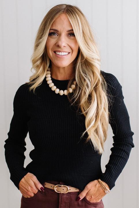 Blonde woman standing in a brightly lit studio with light wood floors and paneled wall behind. Wearing dark pants and a ribbed slim fit long sleeve sweater in black.