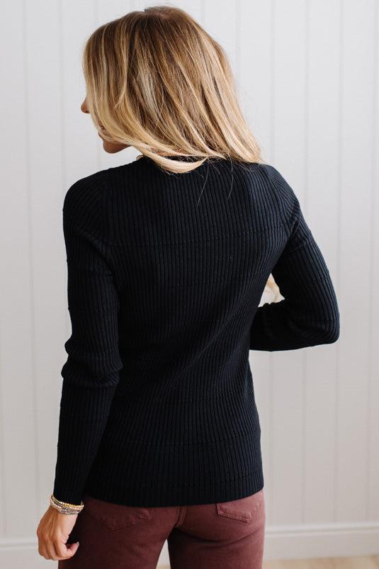 Blonde woman standing in a brightly lit studio with light wood floors and paneled wall behind. Wearing dark pants and a ribbed slim fit long sleeve sweater in black.