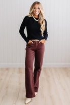 Blonde woman standing in a brightly lit studio with light wood floors and paneled wall behind. Wearing dark pants and a ribbed slim fit long sleeve sweater in black.