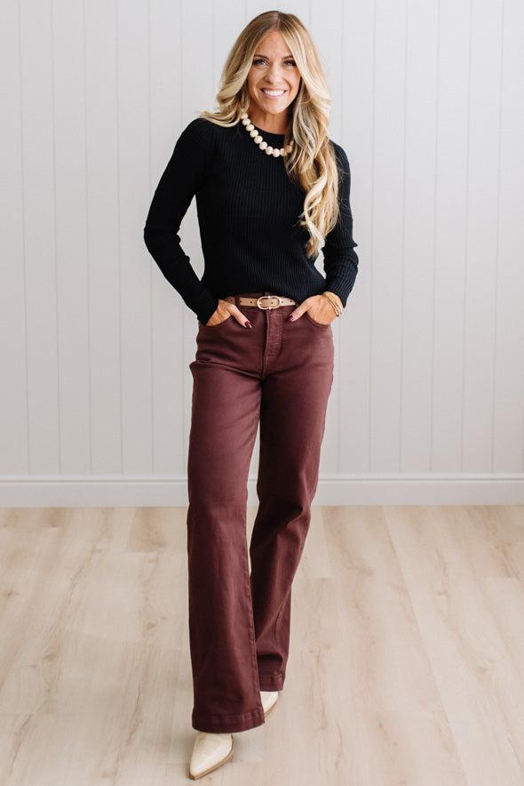 Blonde woman standing in a brightly lit studio with light wood floors and paneled wall behind. Wearing dark pants and a ribbed slim fit long sleeve sweater in black.