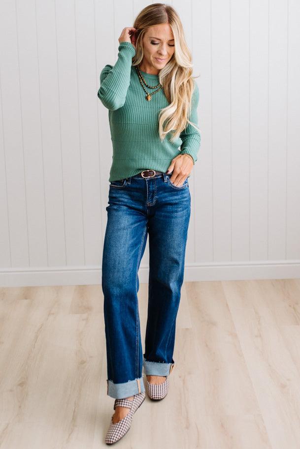 Blonde woman standing in a brightly lit studio with light wood floors and paneled wall behind. Wearing dark pants and a ribbed slim fit long sleeve sweater in green.