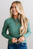 Blonde woman standing in a brightly lit studio with light wood floors and paneled wall behind. Wearing dark pants and a ribbed slim fit long sleeve sweater in green.