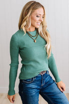 Blonde woman standing in a brightly lit studio with light wood floors and paneled wall behind. Wearing dark pants and a ribbed slim fit long sleeve sweater in green.