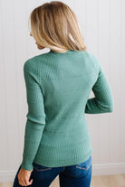 Blonde woman standing in a brightly lit studio with light wood floors and paneled wall behind. Wearing dark pants and a ribbed slim fit long sleeve sweater in green.