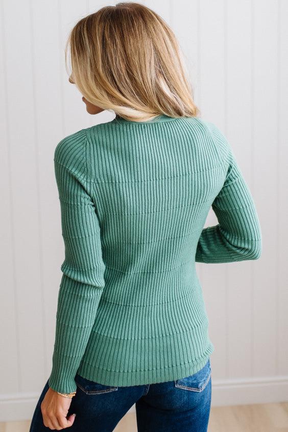 Blonde woman standing in a brightly lit studio with light wood floors and paneled wall behind. Wearing dark pants and a ribbed slim fit long sleeve sweater in green.
