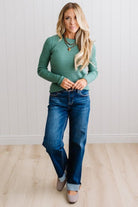 Blonde woman standing in a brightly lit studio with light wood floors and paneled wall behind. Wearing dark pants and a ribbed slim fit long sleeve sweater in green.