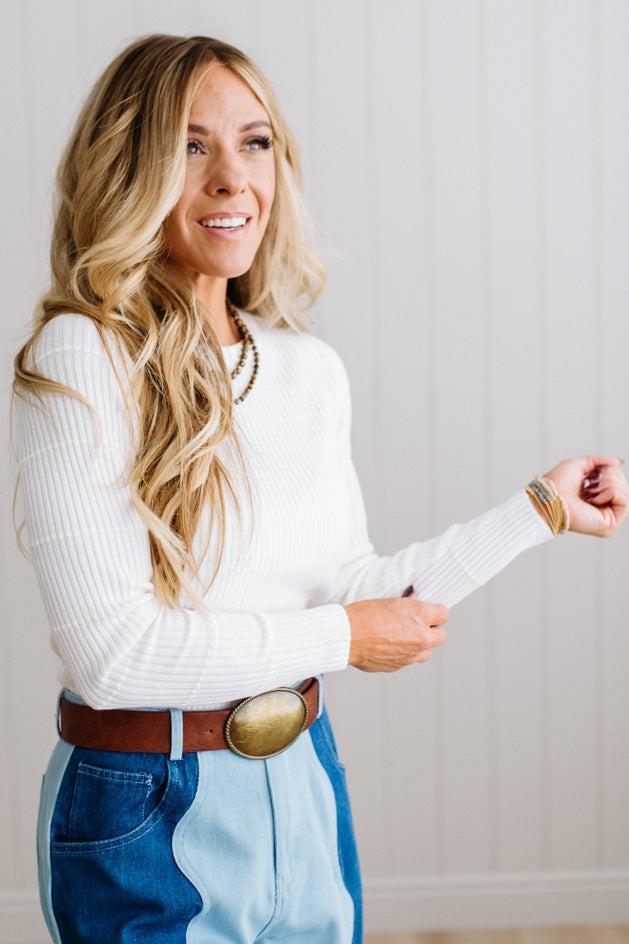 Blonde woman standing in a brightly lit studio with light wood floors and paneled wall behind. Wearing dark pants and a ribbed slim fit long sleeve sweater in white.