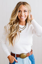 Blonde woman standing in a brightly lit studio with light wood floors and paneled wall behind. Wearing dark pants and a ribbed slim fit long sleeve sweater in white.