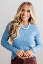 Blonde woman standing in a brightly lit studio with light wood floors and paneled wall behind. Wearing dark pants and a ribbed slim fit long sleeve sweater in blue.