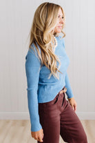 Blonde woman standing in a brightly lit studio with light wood floors and paneled wall behind. Wearing dark pants and a ribbed slim fit long sleeve sweater in blue.