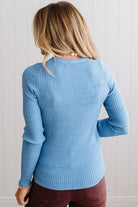 Blonde woman standing in a brightly lit studio with light wood floors and paneled wall behind. Wearing dark pants and a ribbed slim fit long sleeve sweater in blue.