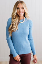 Blonde woman standing in a brightly lit studio with light wood floors and paneled wall behind. Wearing dark pants and a ribbed slim fit long sleeve sweater in blue.