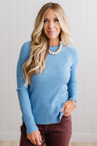 Blonde woman standing in a brightly lit studio with light wood floors and paneled wall behind. Wearing dark pants and a ribbed slim fit long sleeve sweater in blue.