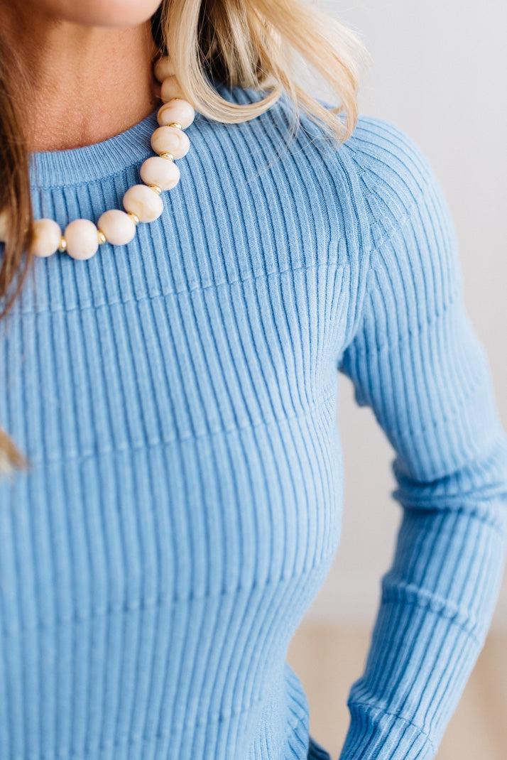 Blonde woman standing in a brightly lit studio with light wood floors and paneled wall behind. Wearing dark pants and a ribbed slim fit long sleeve sweater in blue.