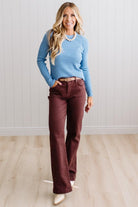 Blonde woman standing in a brightly lit studio with light wood floors and paneled wall behind. Wearing dark pants and a ribbed slim fit long sleeve sweater in blue.