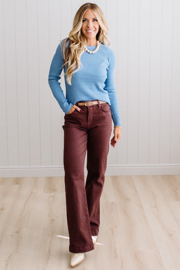 Blonde woman standing in a brightly lit studio with light wood floors and paneled wall behind. Wearing dark pants and a ribbed slim fit long sleeve sweater in blue.