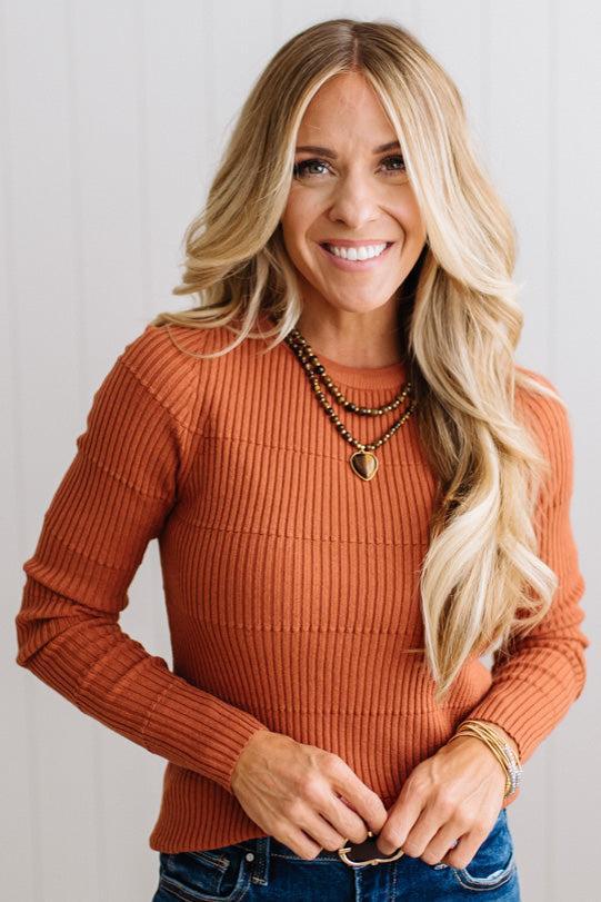 Blonde woman standing in a brightly lit studio with light wood floors and paneled wall behind. Wearing dark pants and a ribbed slim fit long sleeve sweater in rust.