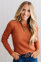Blonde woman standing in a brightly lit studio with light wood floors and paneled wall behind. Wearing dark pants and a ribbed slim fit long sleeve sweater in rust.
