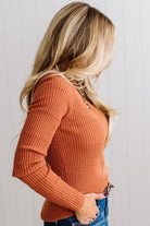Blonde woman standing in a brightly lit studio with light wood floors and paneled wall behind. Wearing dark pants and a ribbed slim fit long sleeve sweater in rust.