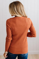 Blonde woman standing in a brightly lit studio with light wood floors and paneled wall behind. Wearing dark pants and a ribbed slim fit long sleeve sweater in rust.