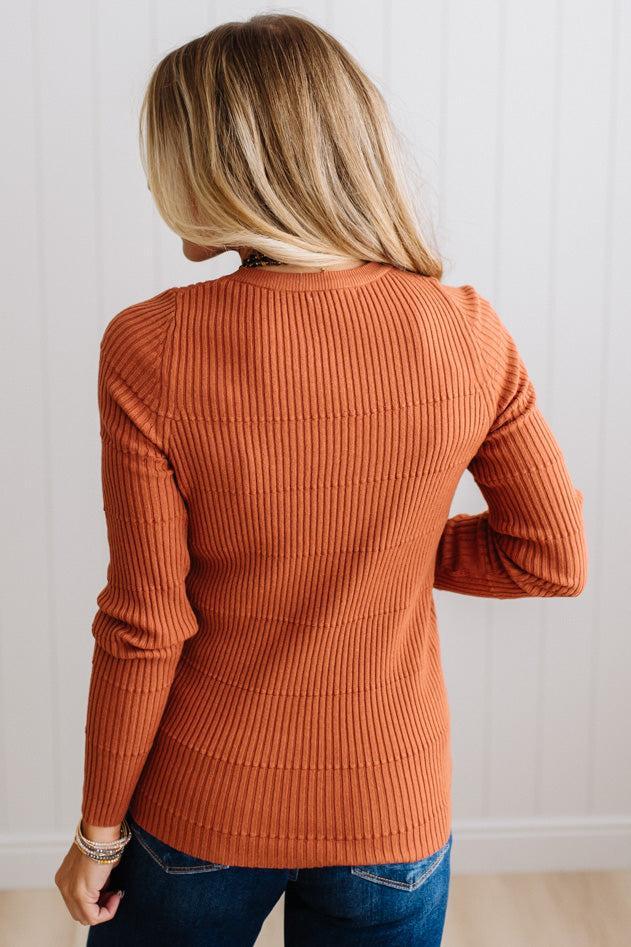 Blonde woman standing in a brightly lit studio with light wood floors and paneled wall behind. Wearing dark pants and a ribbed slim fit long sleeve sweater in rust.