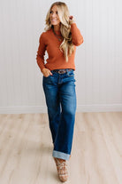 Blonde woman standing in a brightly lit studio with light wood floors and paneled wall behind. Wearing dark pants and a ribbed slim fit long sleeve sweater in rust.